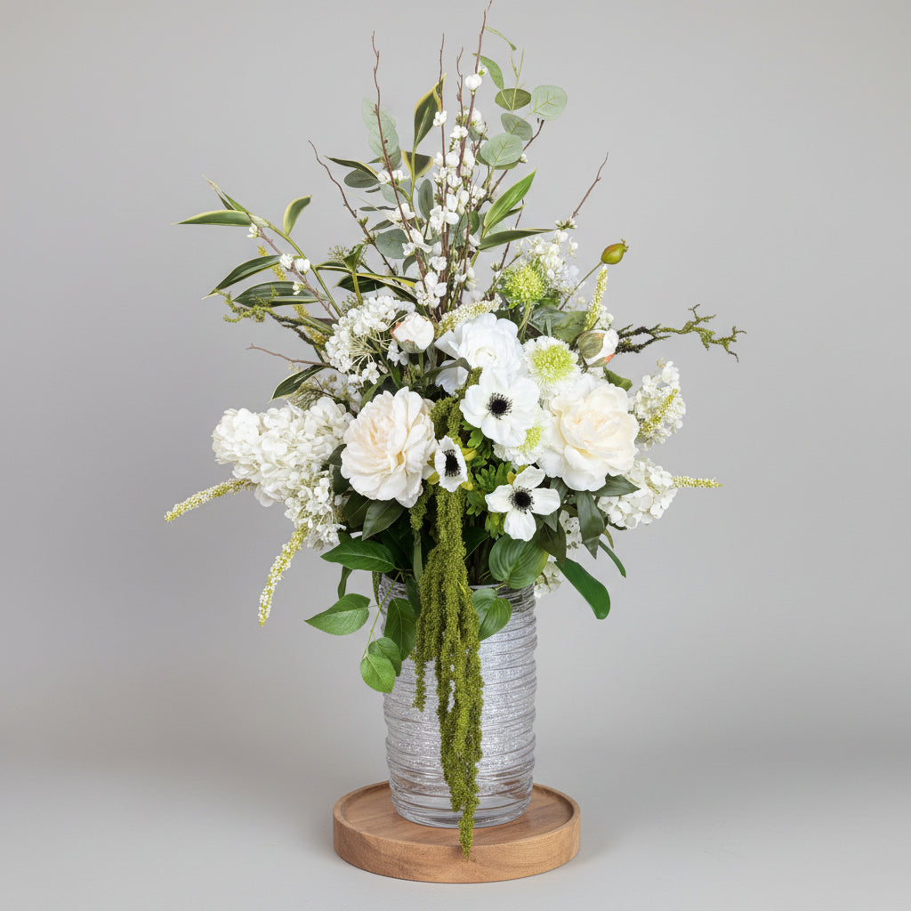 A large floral arrangement featuring white roses, hydrangeas, anemones, and green foliage in a textured white vase on a dark wooden stand, set inside a flower shop.